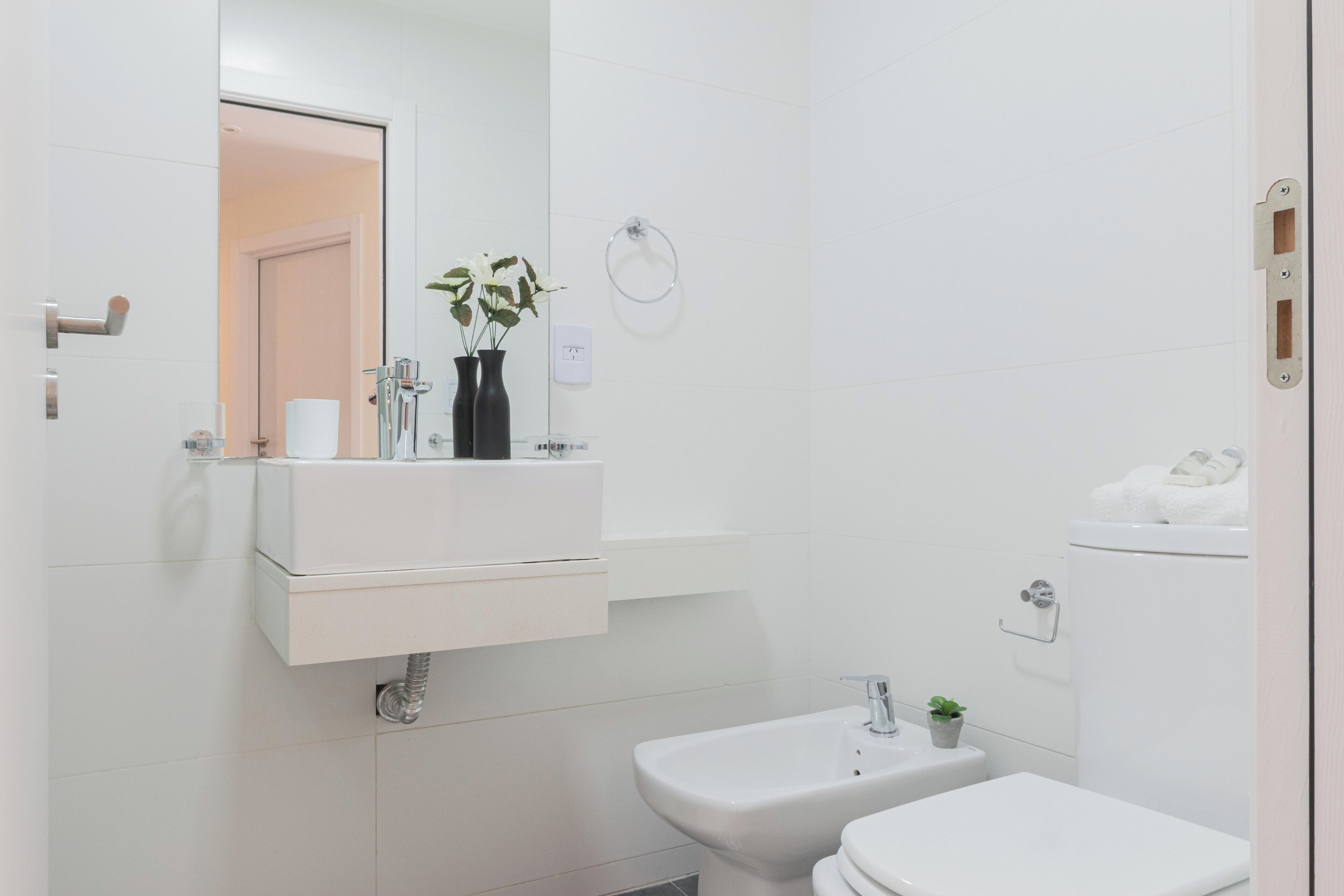 Et moderne badeværelse med en hvid vask, spejl og toilet. Der er to vaser med planter på vaskeskålen.