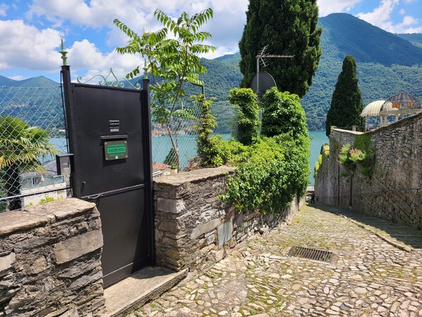 A stone pathway leading to a gate with a 'For Rent' sign, surrounded by lush greenery and a view of a lake and mountains. - Villa "La Gabbianella" Overview in Moltrasio, Como (Moltrasio)