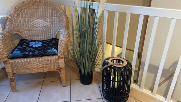 A wicker chair with a floral pillow, a tall potted plant, and a black lantern beside a white railing. - Ferienhof Eckhoff apartment no. 1 (Fehmarn)