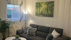 A living room with a sectional sofa, a round coffee table, a floor lamp, a painting of a forest, and a large window with sheer curtains. - Ferienhof Eckhoff apartment no. 1 (Fehmarn)