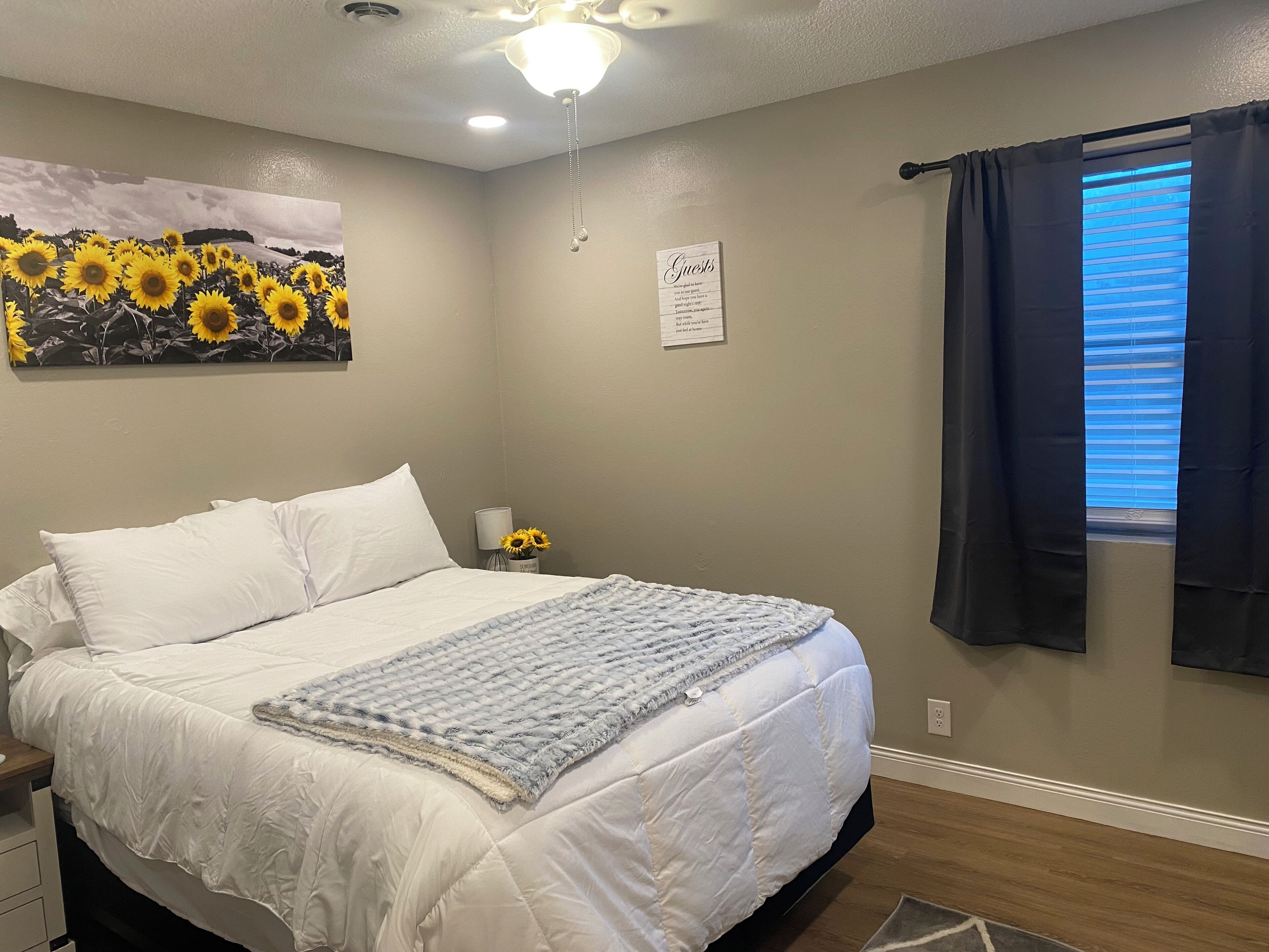 A bedroom with a bed, a nightstand with a lamp and flowers, a ceiling fan, a wall with a 'Guests' sign, and a window with curtains.