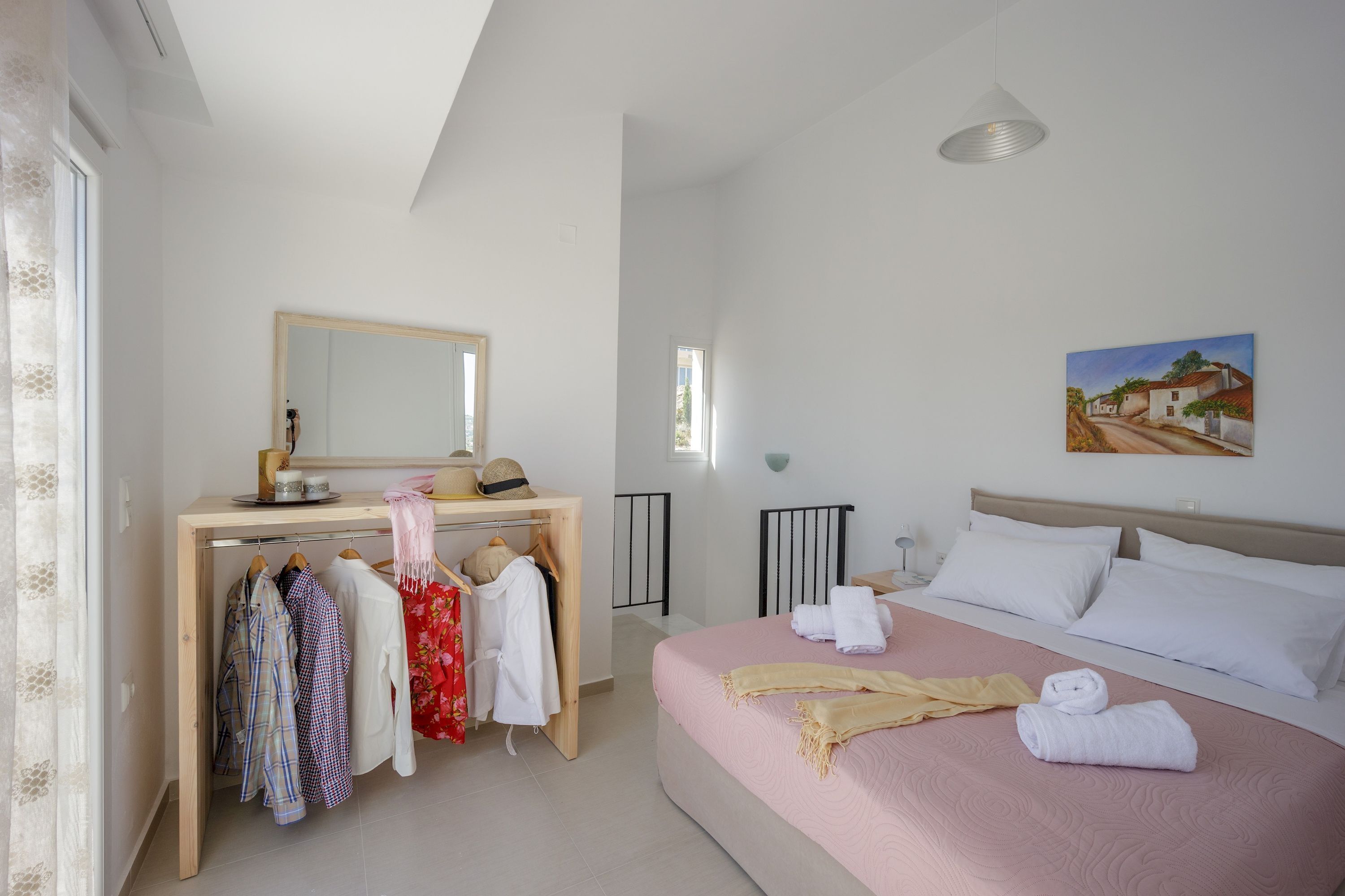A bedroom with a bed, a desk, and a mirror. There are clothes hanging on the wall and a painting on the wall.