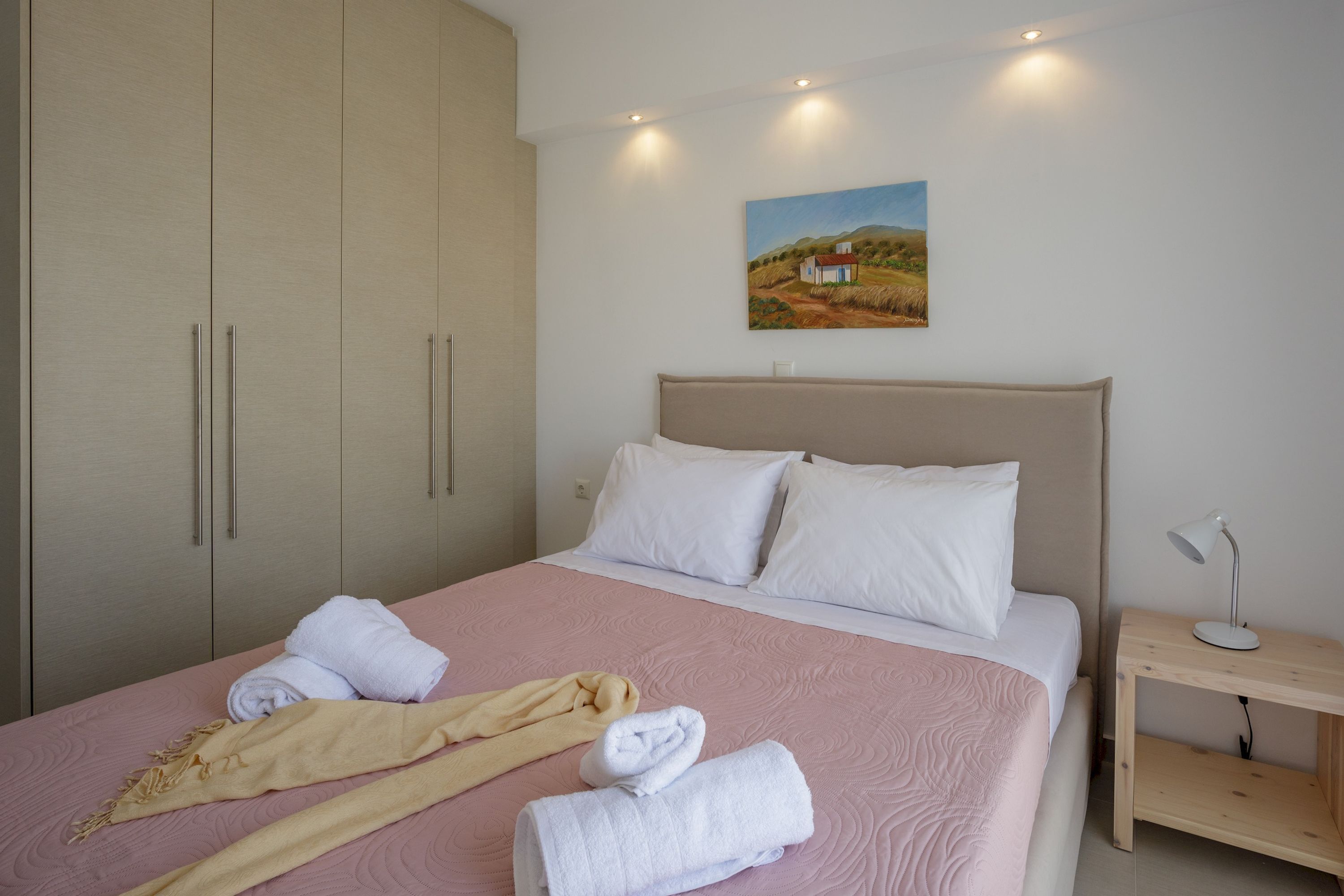 A neatly made bed with white and beige towels, a wooden nightstand with a lamp, and a painting of a rural landscape on the wall.