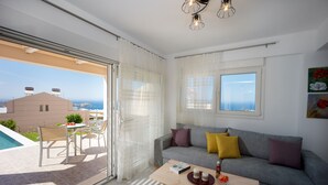 A modern living room with a grey sofa, yellow and purple cushions, a wooden coffee table, and a view of the sea through large windows. - Blue Hills Villas by Estia Purple 1 (Malevizi)