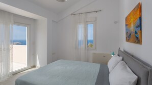 Ein Schlafzimmer mit Bett, einem Fenster mit Vorhängen, einem Bild an der Wand und Blick aufs Meer.