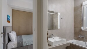 Ein modernes Schlafzimmer mit Bett, Nachttisch, großem Kleiderschrank und einem Badezimmer mit Waschbecken und Badewanne.