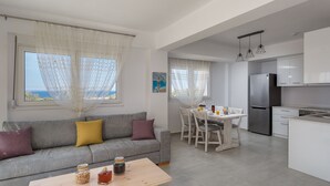 Ein modernes Wohnzimmer mit einem grauen Sofa, einem Holztisch und einem Essbereich mit weißem Tisch und Stühlen. Im Hintergrund ist eine Küche mit weißen Schränken und einem Edelstahl-Kühlschrank zu sehen.