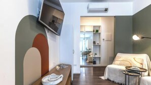 Un salon moderne avec un banc en bois, un canapé et une petite table. Un téléviseur à écran plat est fixé au mur, et un appareil de climatisation est installé juste au-dessus.