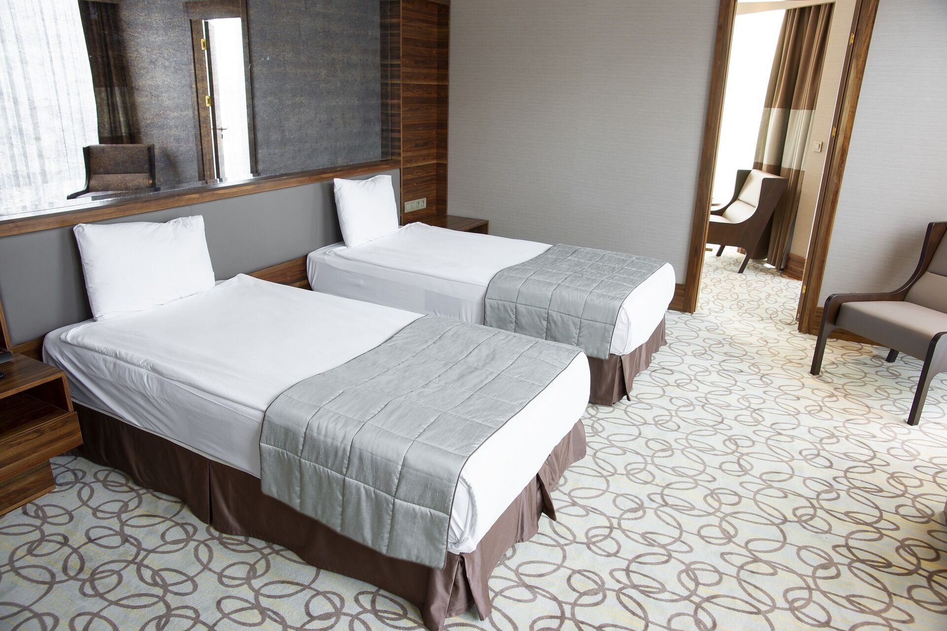 A hotel room with two beds, a wooden headboard, a mirror, a chair, and a patterned carpet.