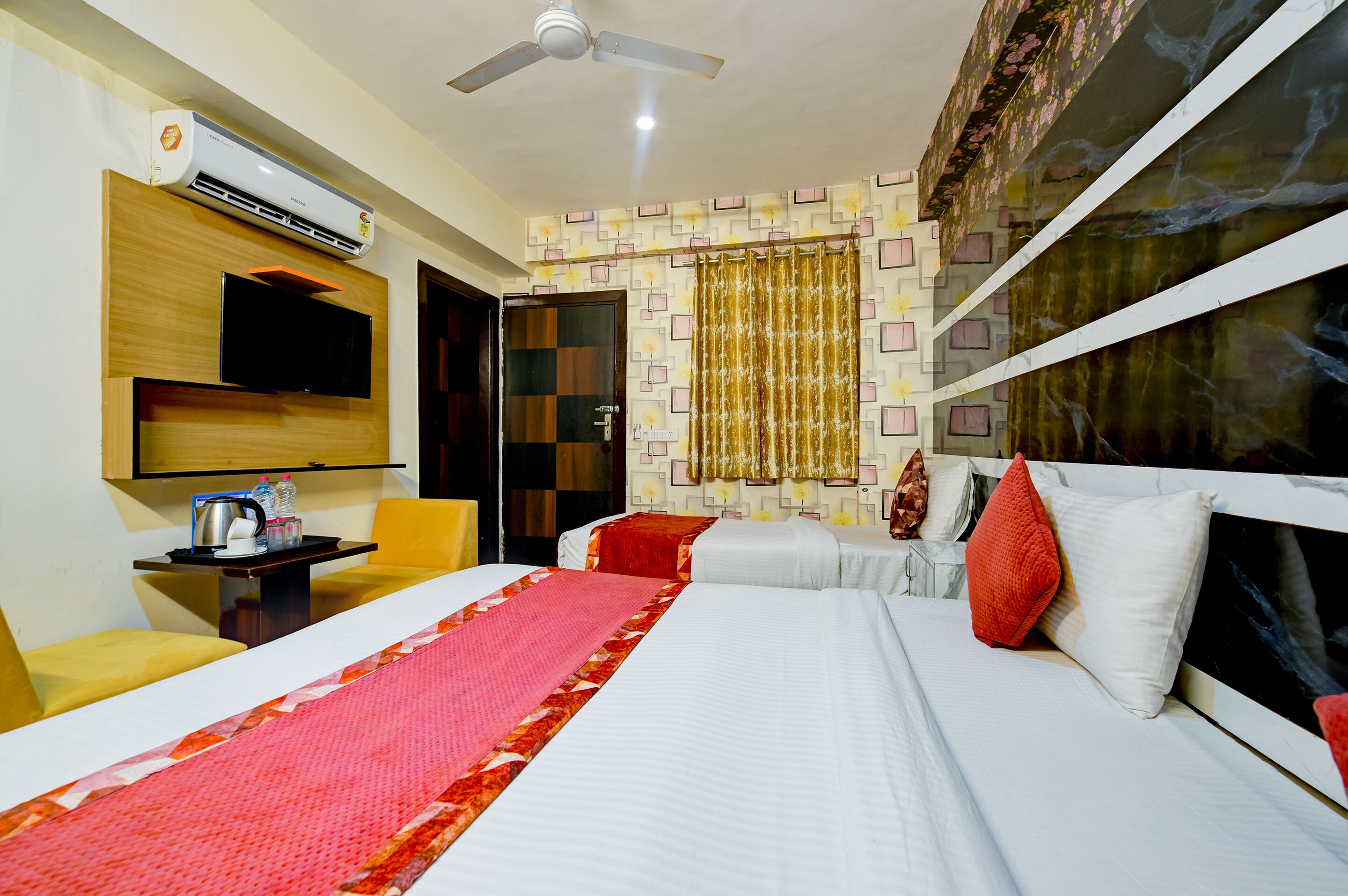 A hotel room with a bed, a television, a ceiling fan, a small table with a kettle, and a window with curtains.