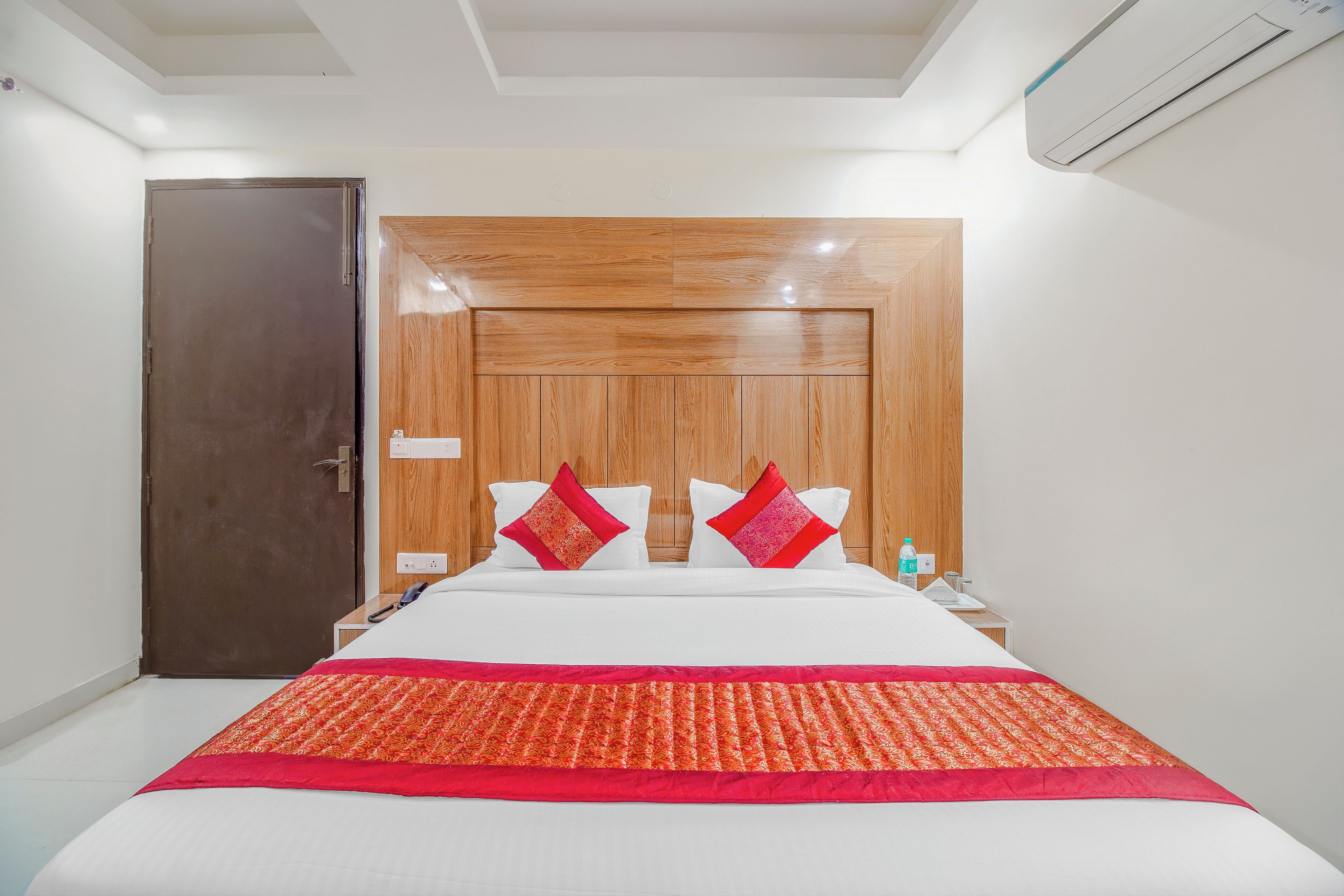 Une chambre d’hôtel avec une tête de lit en bois, un lit recouvert de draps blancs et agrémenté de touches de rouge, une porte et une table de chevet sur laquelle se trouve une bouteille.