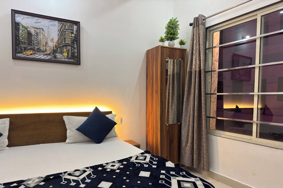 A hotel room with a bed, a nightstand, a wooden wardrobe, a window with curtains, and a painting on the wall.