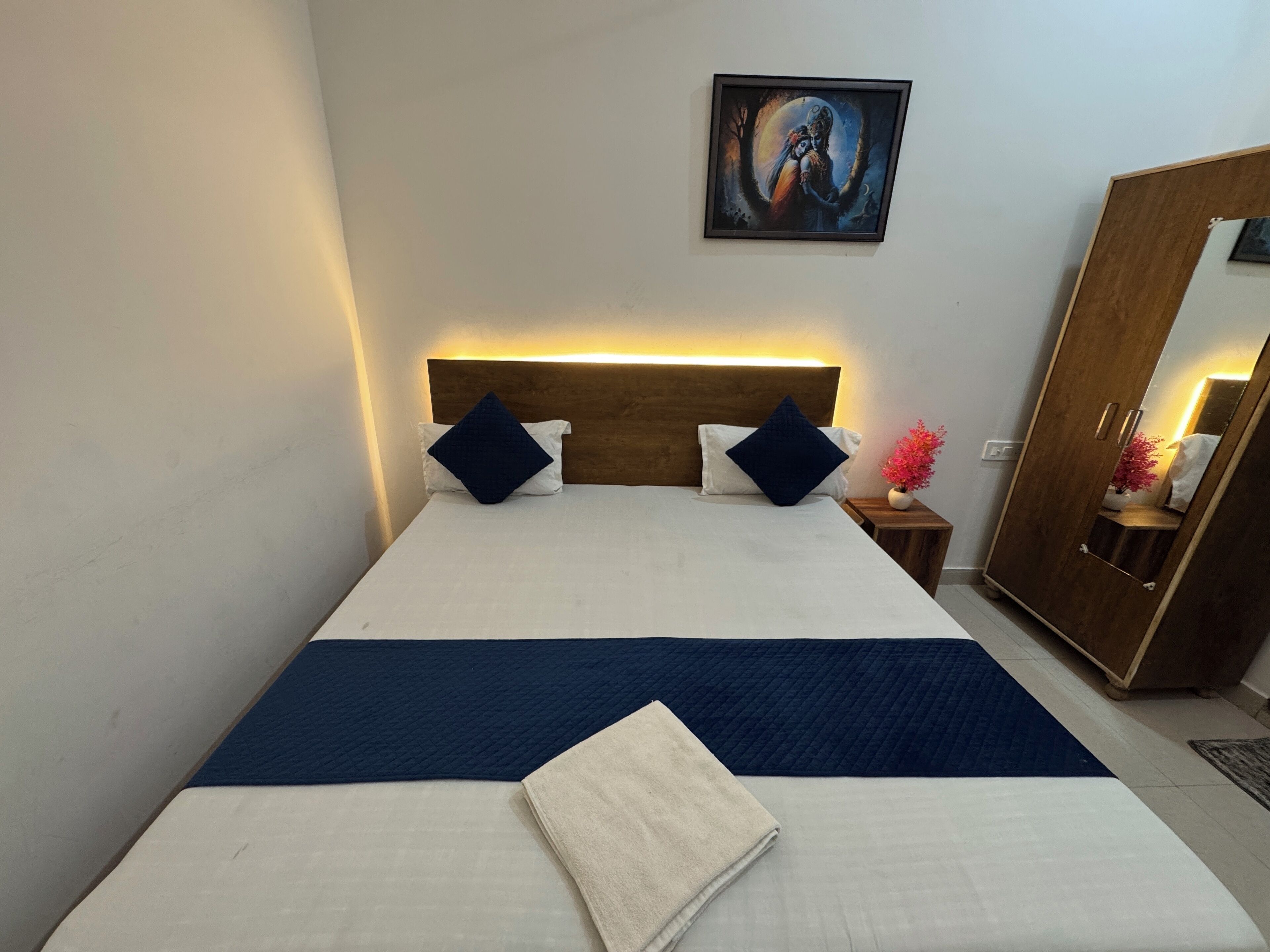 A neatly made bed with a blue and white bedspread, two dark blue pillows, and a white towel on the bed. There is a wooden headboard, a bedside table with a vase of flowers, and a large mirror on the wall.