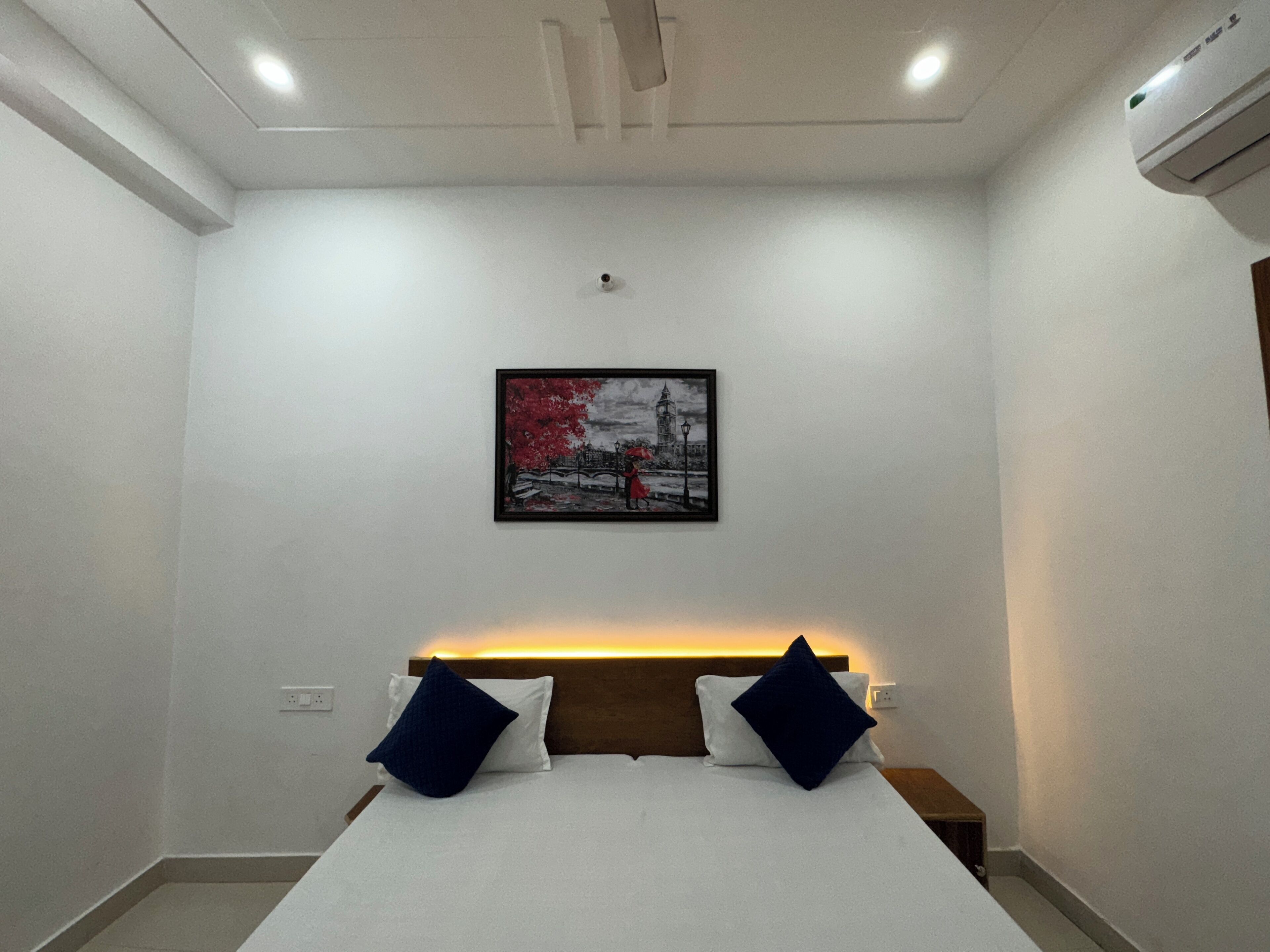 A hotel room with a bed, a nightstand, a wall-mounted air conditioner, and a framed picture on the wall.