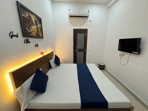 A neatly made bed with a headboard, a wall-mounted TV, an air conditioning unit, and a framed picture above the bed. - Shree Radha Vallabh Palace- 800 m from prem mandir (Vrindavan)