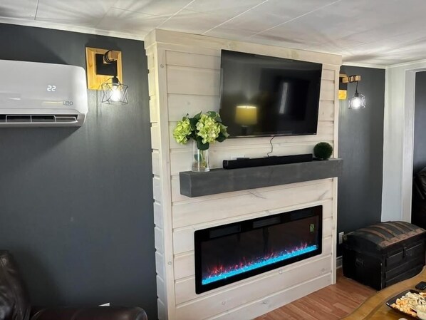 A living room with a flat-screen TV mounted above a fireplace, a wall-mounted air conditioner, and a small table with snacks.