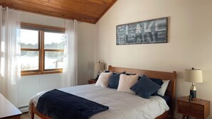 A bedroom with a wooden ceiling, a bed with a striped bedspread, multiple pillows, a nightstand with a lamp, and a window with curtains.