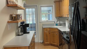 A modern kitchen with wooden cabinets, a black refrigerator, and a built-in oven. There are wooden shelves above the counter and a window with blinds.