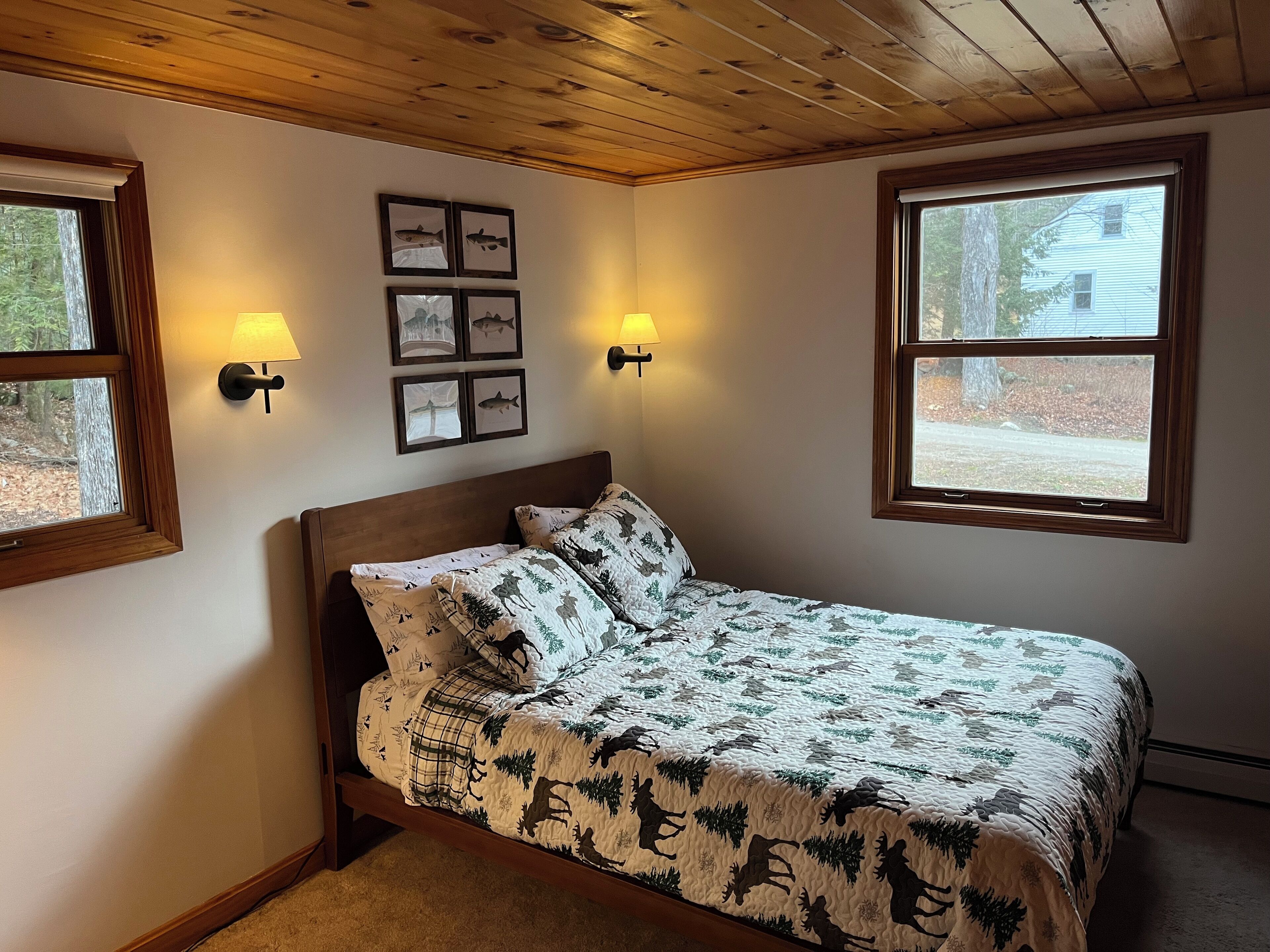Un dormitorio con cama de madera, dos lámparas fijadas a la pared, cuadros enmarcados y ventanas amplias.