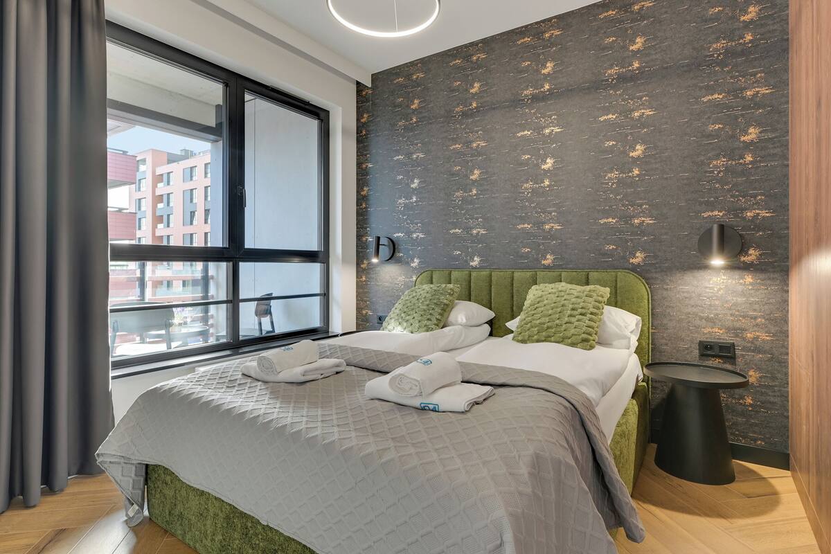 Ett modernt hotellrum med ett stort fönster, en grön klädd sänggavel, ett runt svart sidobord och en säng med prydligt hopvikta handdukar.