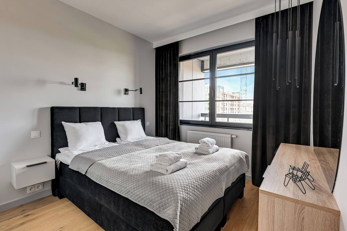 Ett modernt hotellrum med en stor säng, ett nattduksbord, ett fönster med gardiner och utsikt över byggnader.