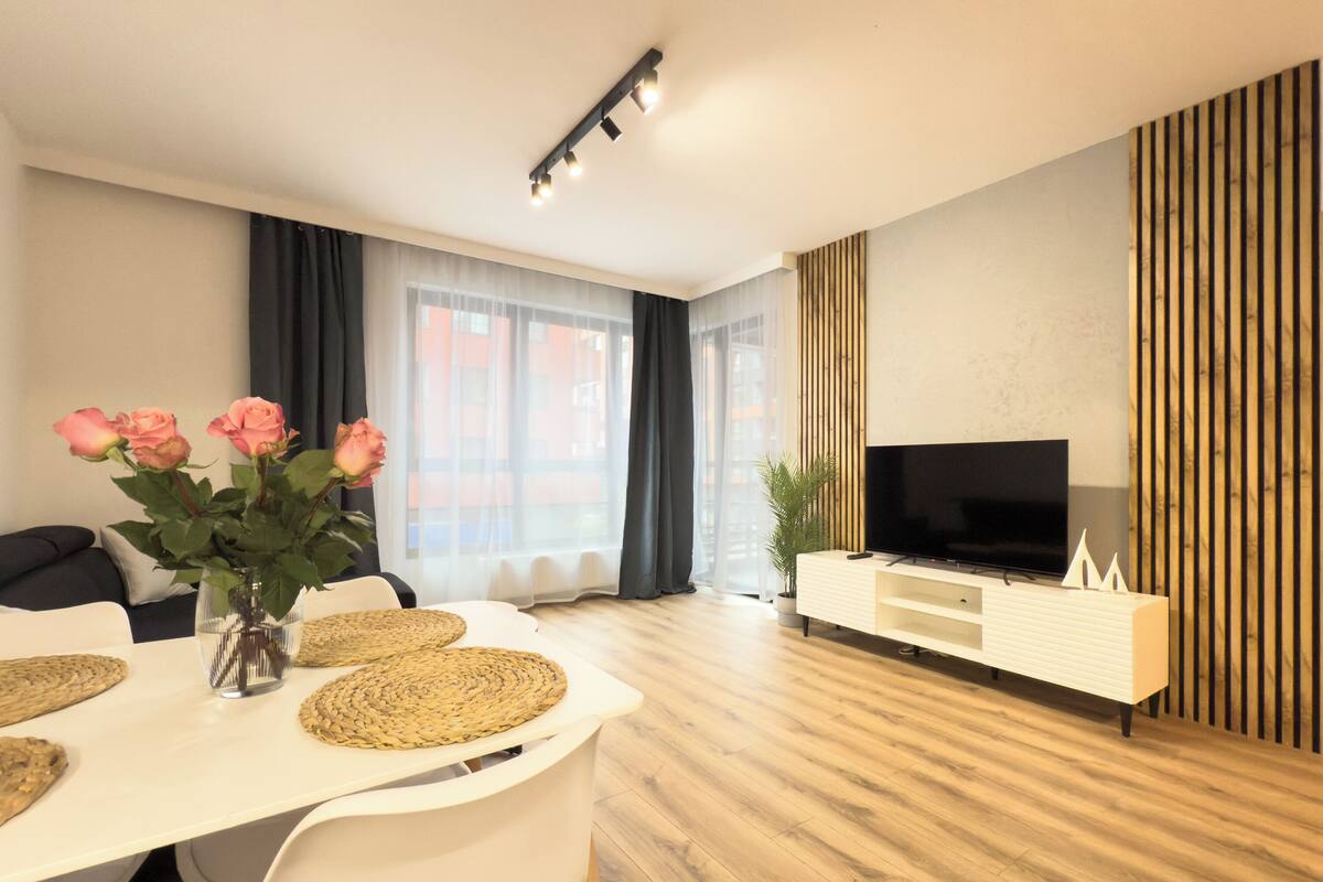 Ett modernt vardagsrum med en matplats, med en TV, en vit soffa, ett matbord med vävda underlägg och en vas med rosa rosor.