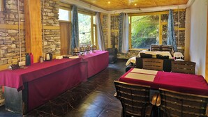 A dining area with a long table set for a meal, stone walls, wooden ceiling, and large windows with curtains. - Crystal Mountain Jibhi (Banjar)