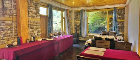 A dining area with a long table set for a meal, stone walls, wooden ceiling, and large windows with curtains.