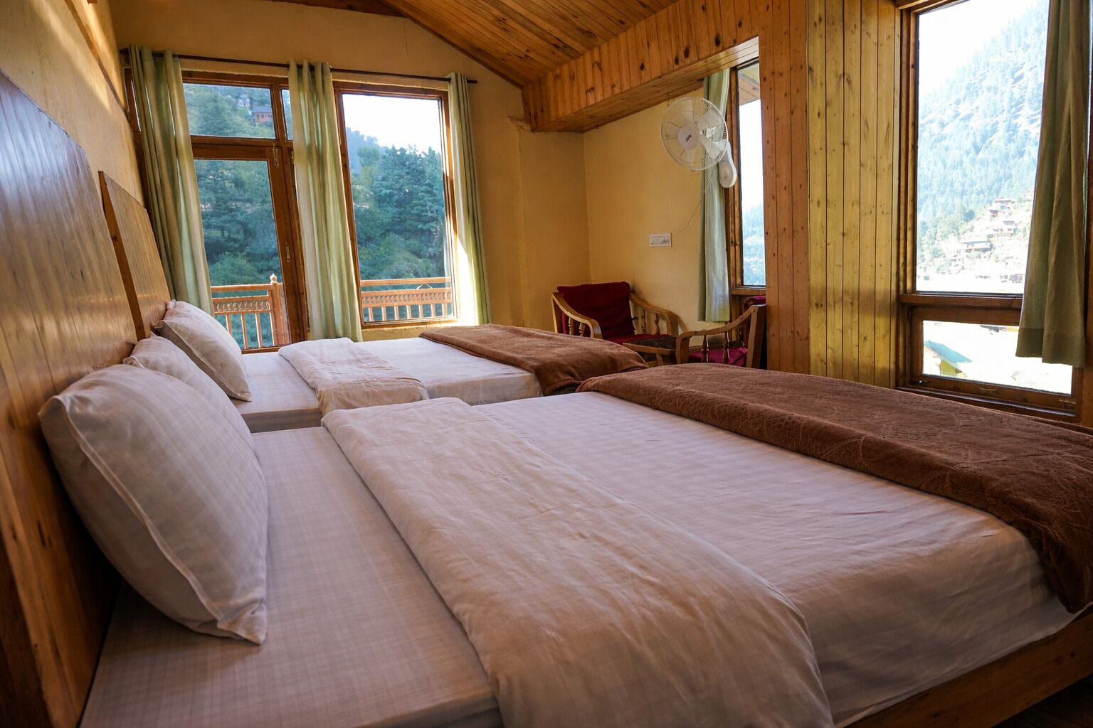 A room with three beds, a chair, a fan, and large windows with curtains.