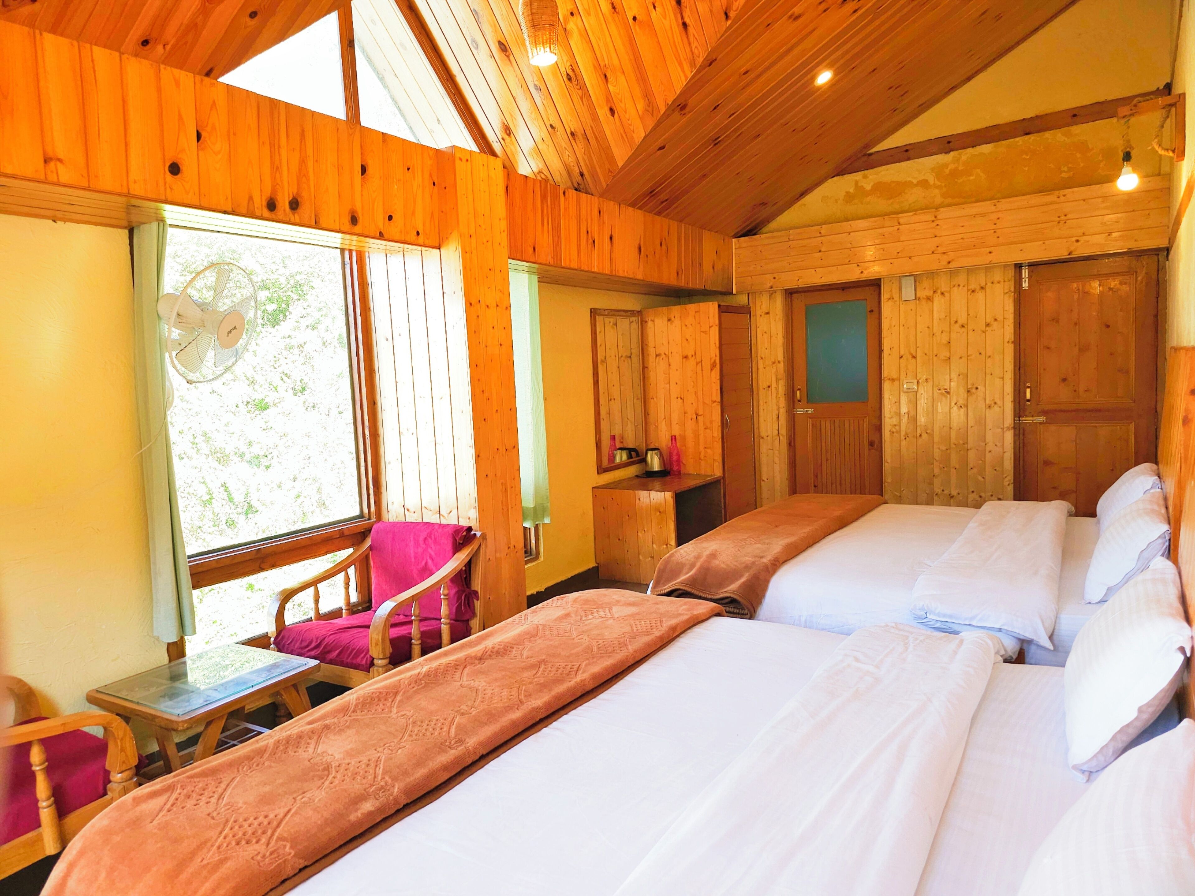 A wooden cabin room with two beds, a table, a chair, and a fan.