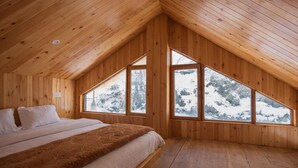 A wooden cabin bedroom with a bed, a window, and a view of a snowy landscape. - Crystal Mountain Jibhi (Banjar)