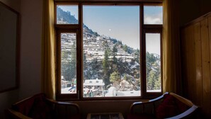 A room with a large window overlooking a mountainous landscape. There are two wooden chairs and a small table in the foreground. - Crystal Mountain Jibhi (Banjar)