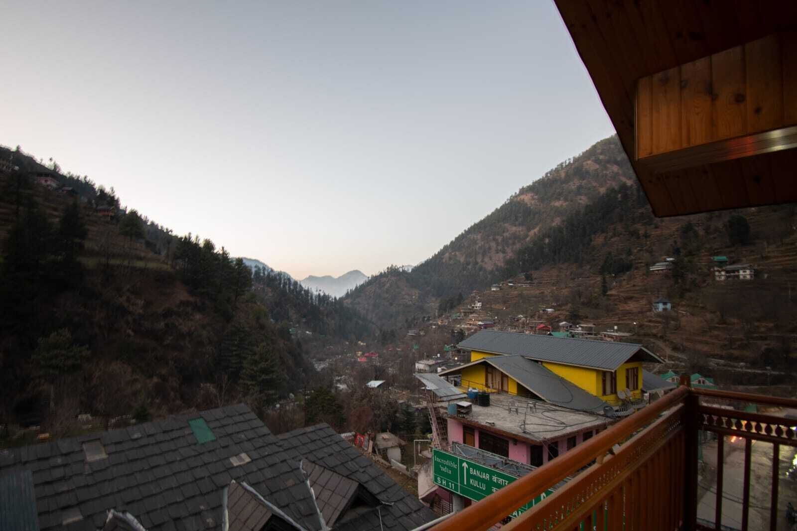 A mountainous landscape with a yellow building and a green sign indicating directions to Banjar and Kullu.