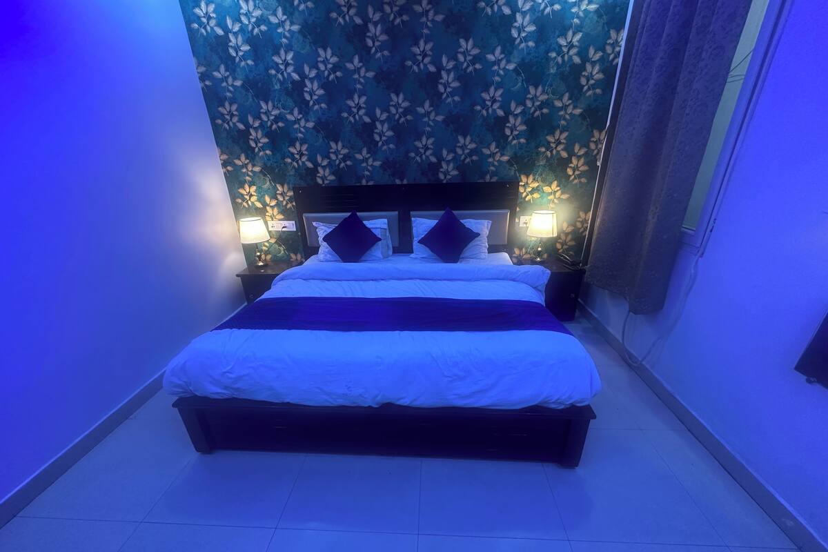 Una cama bien hecha con sábanas blancas y almohadas azul oscuro, flanqueada por dos mesitas de noche con lámparas, en una habitación con papel tapiz floral y suelo de baldosas.