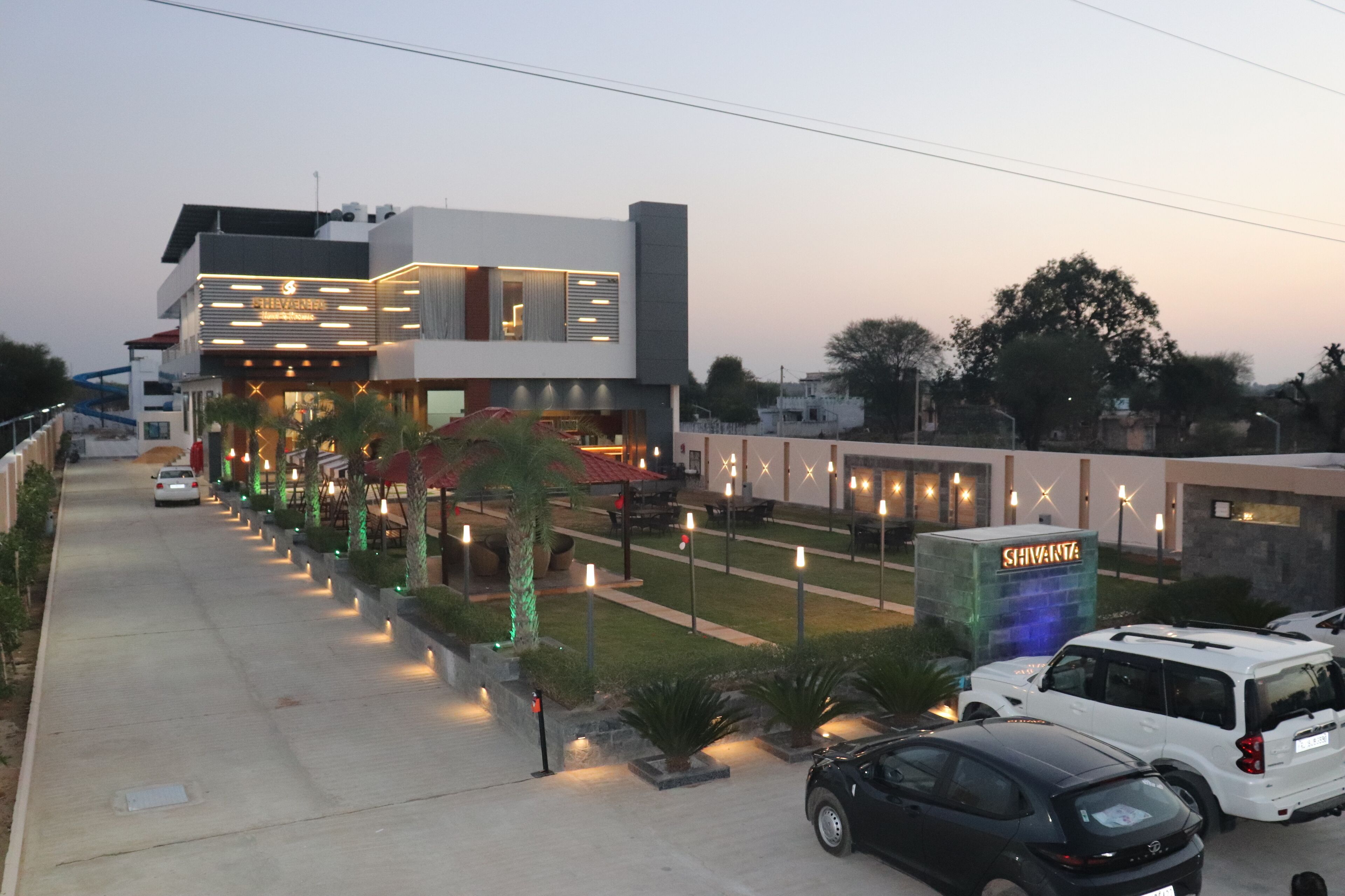 A modern hotel with a well-lit entrance, a parking area, and a visible sign for 'Shivakant'.