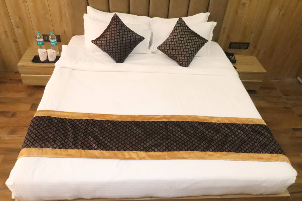 A neatly made bed with white linens and two patterned pillows, a wooden headboard, and a bedside table with a bottle and glasses.