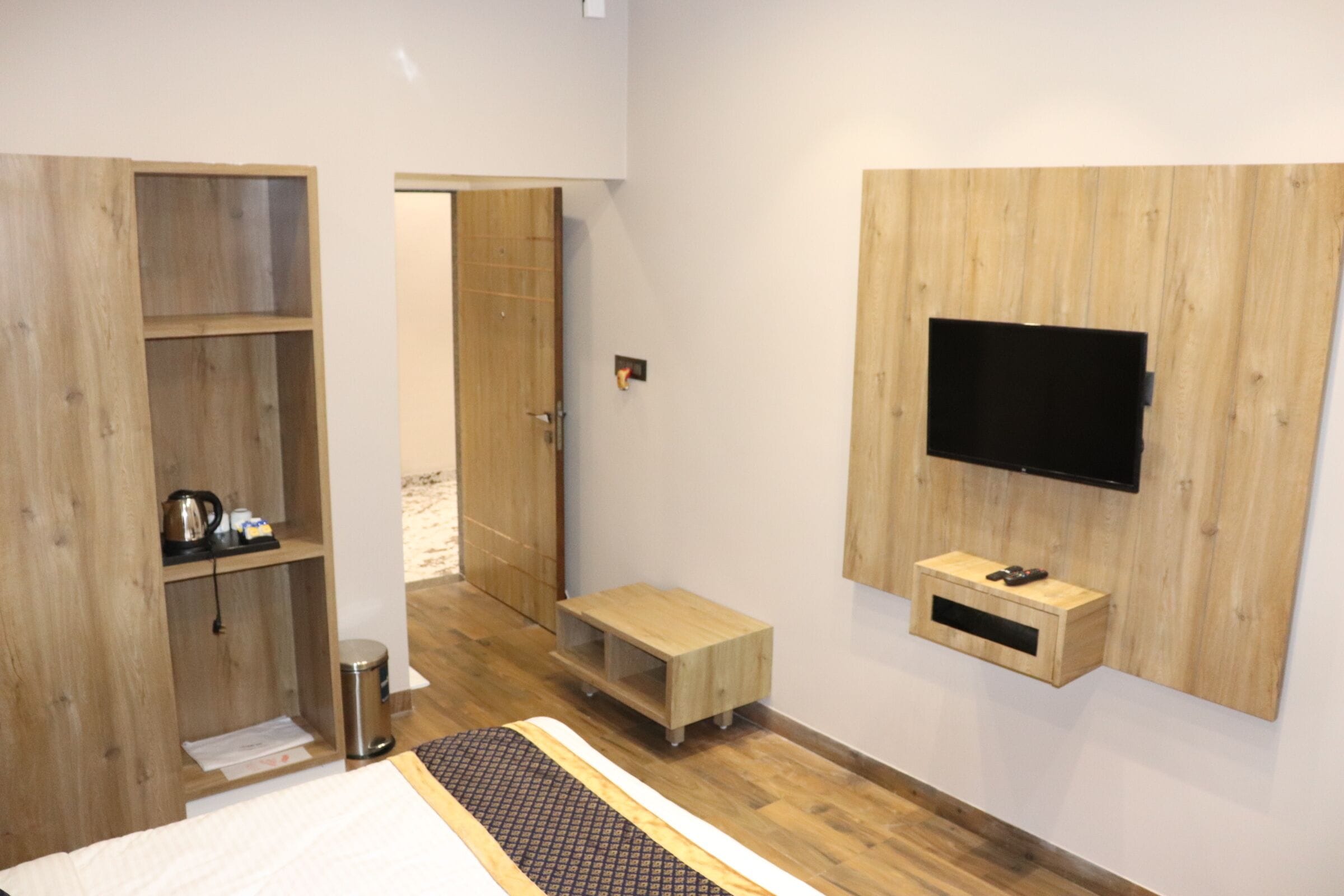 A modern hotel room with a wooden wardrobe, a small seating area, a television mounted on the wall, and a visible door leading to another room.