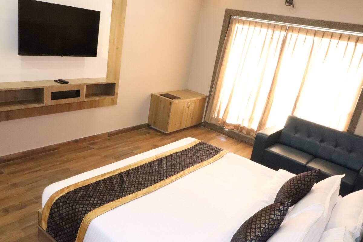 A hotel room with a large bed, a TV mounted on the wall, a wooden cabinet, a small wooden stool, and a dark-colored sofa.