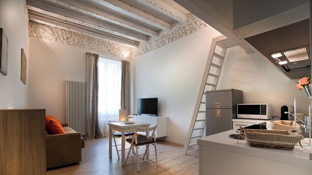 A modern apartment with a loft-style ceiling, a dining area, a kitchen with a refrigerator and microwave, and a TV. - STUDIO BIJOUX (Verona)
