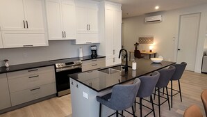 A modern kitchen with white cabinets, a black countertop, and a central island with bar stools. - Spacious 3 bedroom condo in the heart of Mount Orford's nature (Orford)