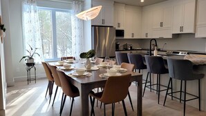 A modern kitchen with white cabinets, a stainless steel refrigerator, and a dining area set for six with a table, chairs, and a vase of flowers. - Spacious 3 bedroom condo in the heart of Mount Orford's nature (Orford)