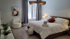 A bedroom with a large bed, a chair, a bedside table with a lamp, a window with curtains, and a plant. - Spacious 3 bedroom condo in the heart of Mount Orford's nature (Orford)