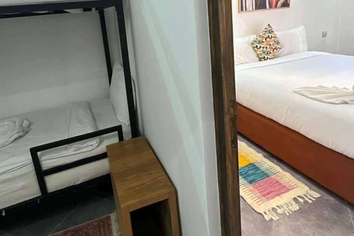 Une pièce avec des lits superposés, un petit tabouret en bois, un tapis coloré et un tableau accroché au mur.