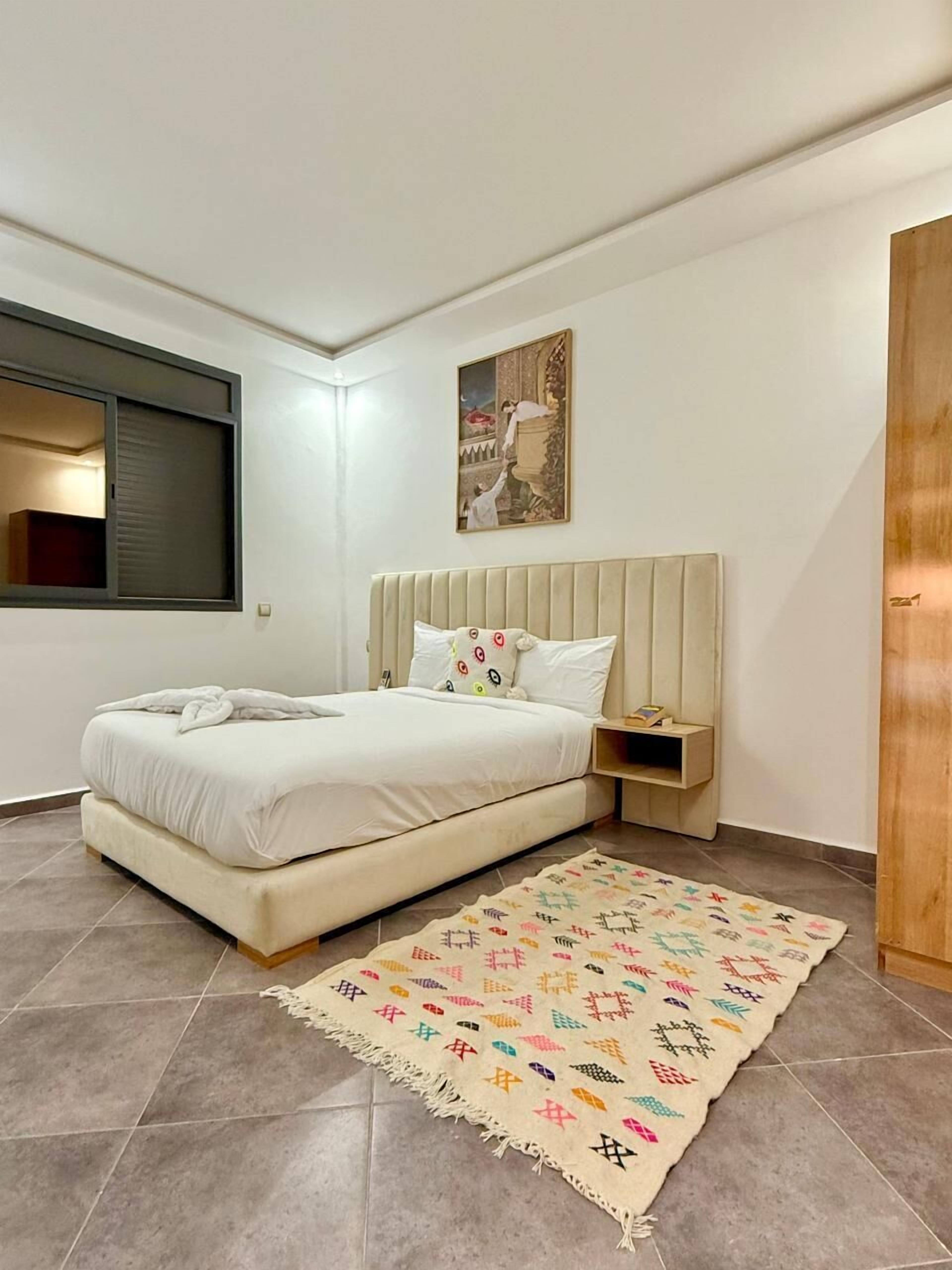 A bedroom with a bed, a bedside table, a rug, a window, and a painting on the wall.