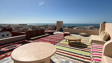 Terraço no telhado com mesa redonda, tapetes coloridos e vista para o mar e para a paisagem urbana.