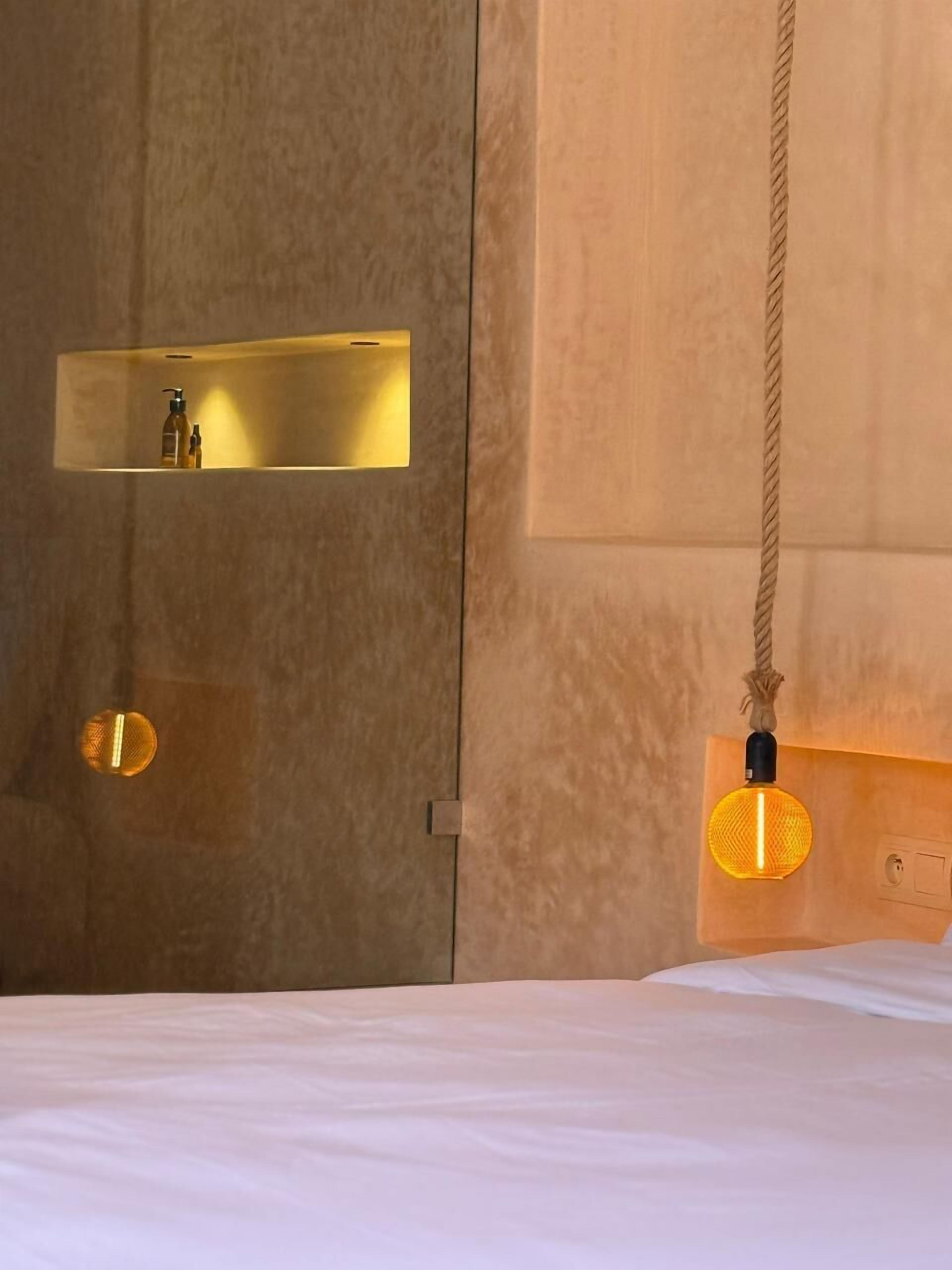 A hotel room with a bed, a wall-mounted shelf with toiletries, a round light fixture, and a rope hanging from the ceiling.