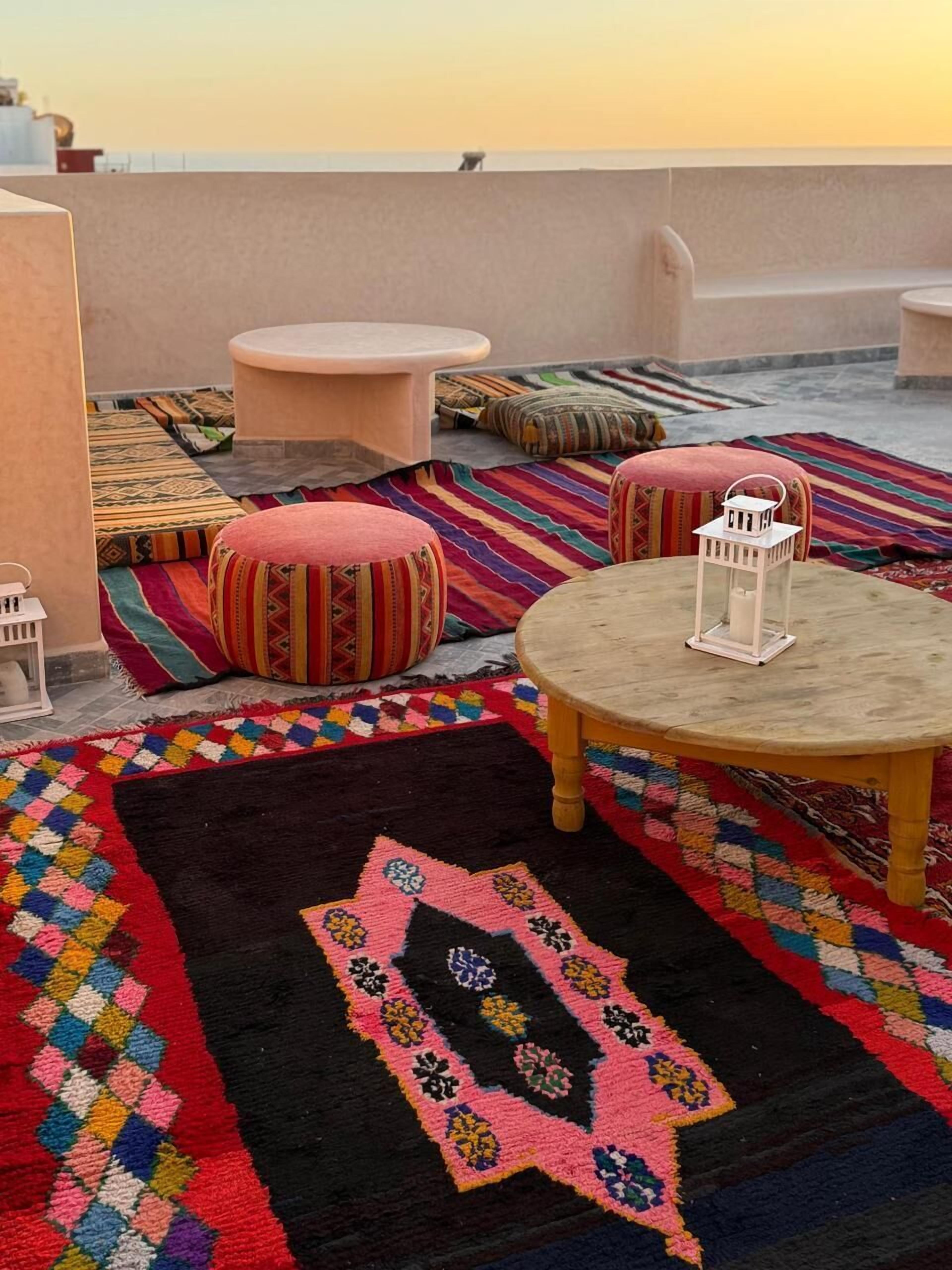A rooftop terrace with colorful rugs, round tables, and ottomans.