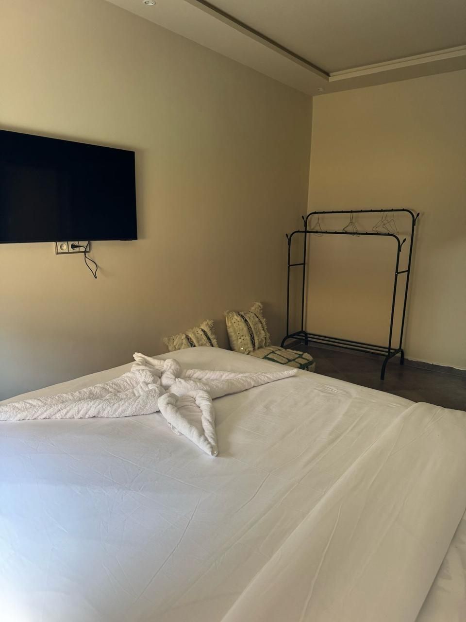 A hotel room with a neatly made bed, a TV mounted on the wall, and a metal rack against the wall.