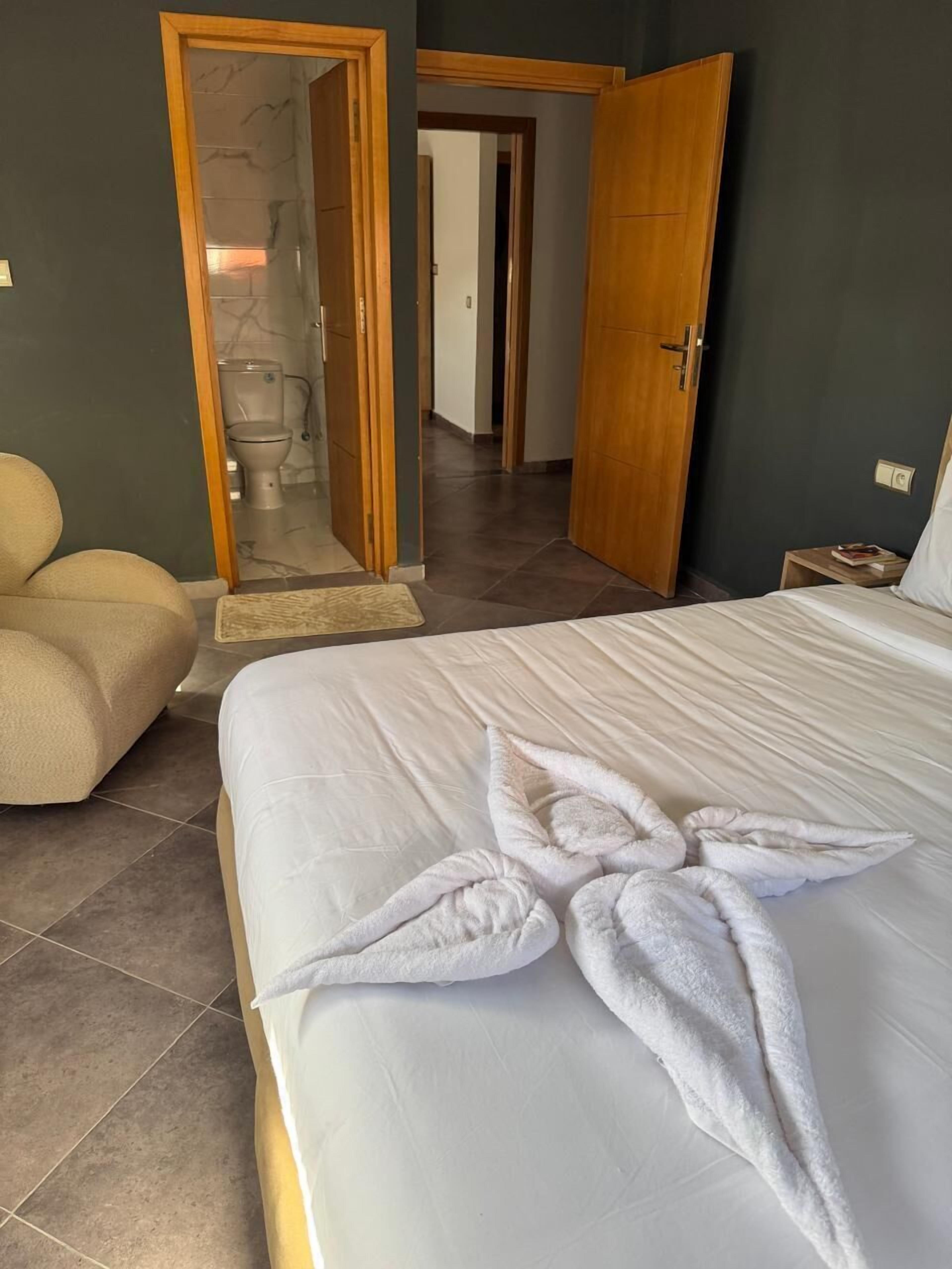 Habitación de hotel con una cama, una puerta de madera, un baño con inodoro y un sillón beige.