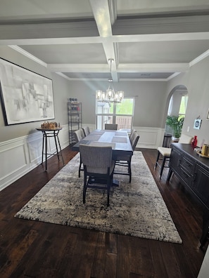 A dining room with a long table, chairs, a chandelier, a large abstract painting, and a view through a window.